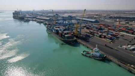 An aerial photograph of a bustling port terminal in Basra, showing a large container vessel docked at blue cranes, a small tugboat in the turquoise water, and stacks of shipping containers on the dock, visually representing the vital maritime infrastructure that facilitates Iraqi Oil Exports, while highlighting the complex geopolitical risks of these economic flows indirectly benefiting Iran.