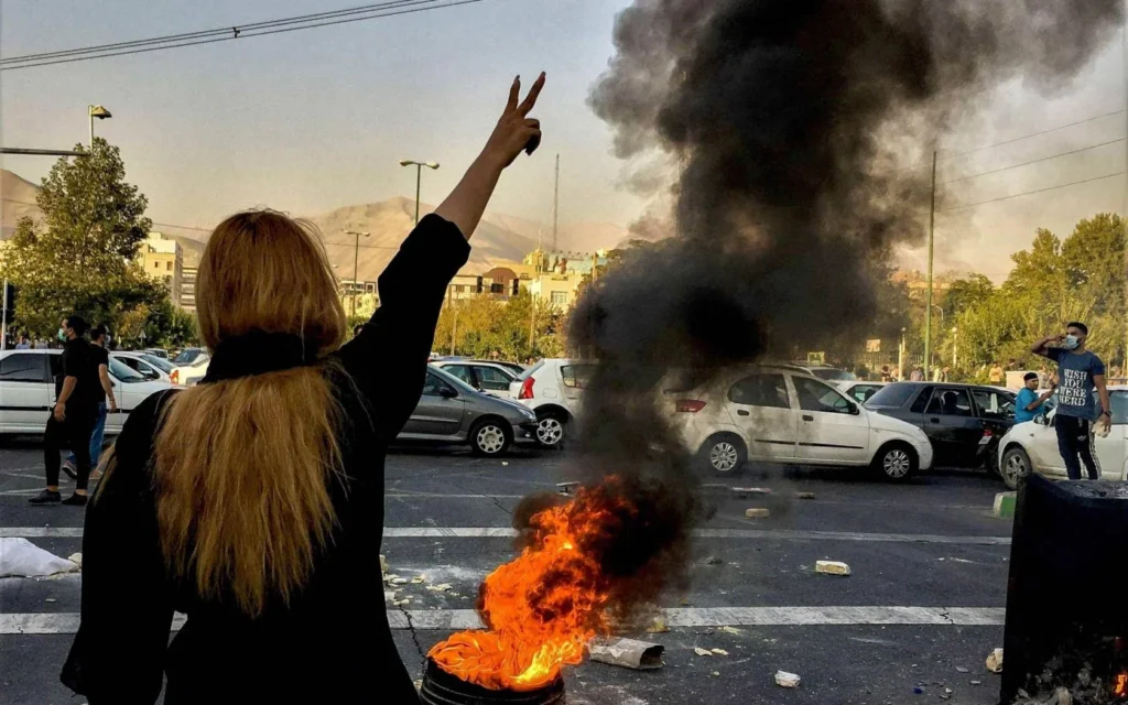 A woman flashes a victory sign beside a street fire as Iranian public opinion backs strikes for regime change.