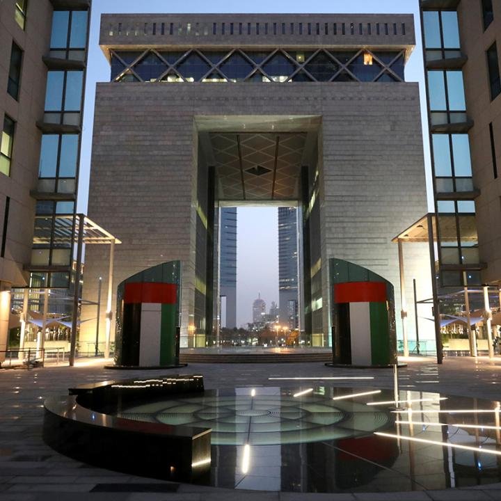 تحریمهای امارات هدف عملیات بانکی سایه ایران قرار گرفت A dusk photograph of a modern stone-and-glass financial gate, flanked by towers and cylindrical pillars painted with the UAE flag colors that reflect in a central glass pool, symbolizing the heavily regulated financial hub where the stringent UAE Sanctions are being enforced to target and dismantle Iran's shadow banking operations.
