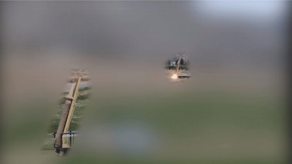 A distant, heavily blurred screen capture from an IRGC video showing a ballistic missile elevated on a mobile launcher in an open grassy field, alongside another missile firing a bright orange exhaust plume into the air, capturing the persistent military brinkmanship that defines the US-Iran Stalemate as both nations navigate a constrained, limited strategic endgame without seeking full-scale escalation.