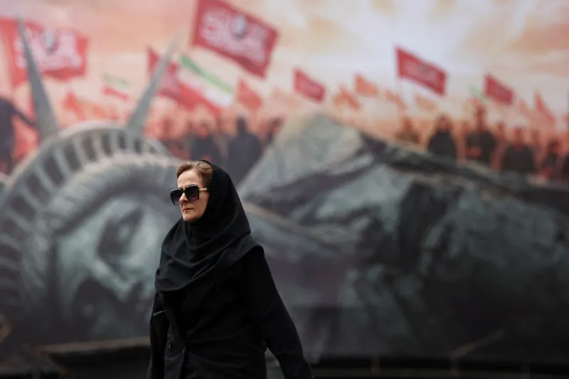Maximum Pressure Fails to Break Iran’s Resistance A woman wearing a black chador and sunglasses walks in the foreground against a blurred anti-American billboard in Tehran depicting a menacing Statue of Liberty, a crowd, and Iranian flags, symbolizing the enduring defiance of daily life that renders the external Maximum Pressure strategy ineffective in breaking Iran's resistance.