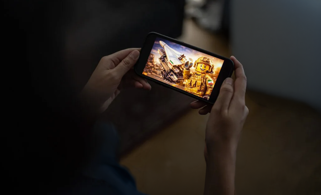 A close-up photograph of hands holding a smartphone displaying a Lego-style animation of an Iranian soldier and a missile, visually representing the viral digital propaganda clips from "Explosive Media" that are central to the Iran Embassy Memes campaign, reshaping global wartime narratives through pro-government imagery.