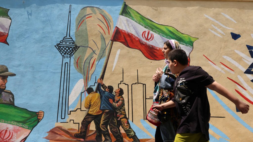 تحریمهای روز D ایران، خطرات درگیری برای خلیج فارس را افزایش میدهد A vibrant street mural in Tehran featuring the Milad Tower and men raising the Iranian flag, with pedestrians walking in the foreground, capturing the defiant public nationalism that heightens regional instability as Iran D-Day sanctions raise severe conflict risks for the Gulf states.