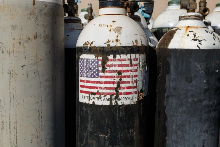 US-Africa Engagement Falters Amid Iran War Distraction A close-up photograph of a highly rusted gas cylinder bearing a peeling and damaged American flag sticker that reads "GIFT FROM THE AMERICAN PEOPLE", set against similar weathered tanks, symbolizing the decaying footprint of humanitarian assistance that underscores how US-Africa Engagement falters amid the strategic distraction of the Iran war.