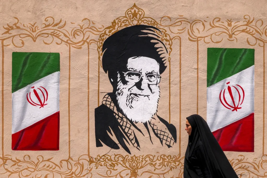 Iran Regime Change Drives Tehran’s Overt Escalation A mural of Ayatollah Khomeini framed by two Iranian flags, with a woman in a black chador walking past, symbolizing the foundational ideology Tehran is fiercely defending against external pressures for Iran Regime Change.