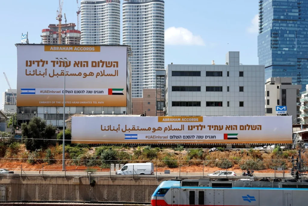 "Large billboards in Tel Aviv celebrating the Abraham Accords and the UAE Embassy, displaying Israeli and Emirati flags and the hashtag #UAEinIsrael, set against a backdrop of modern skyscrapers and a passing train, visually representing the normalization-era framework that current US Middle East interests are now strategically outgrowing."