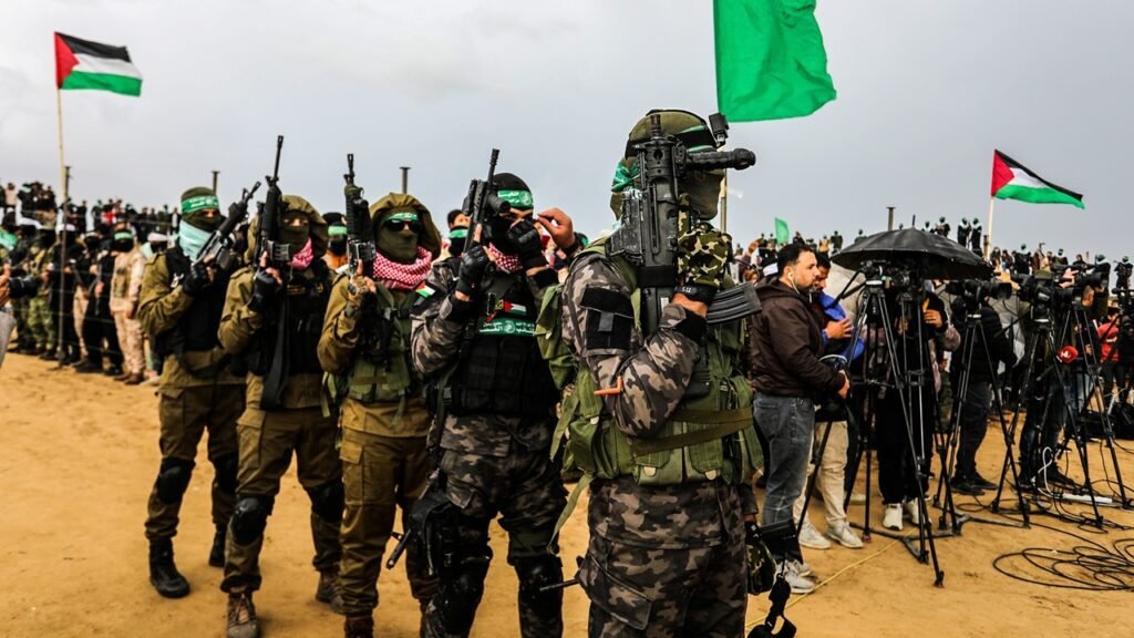 Hamas Intransigence Blocks Gaza Disarmament Roadmap A line of masked Hamas fighters in camouflage tactical gear holding assault rifles, wearing green headbands and checkered keffiyehs, standing defiantly before a crowd of journalists and media cameras under Palestinian and Hamas flags, visually embodying the Hamas Intransigence that continues to block the Gaza Disarmament Roadmap.