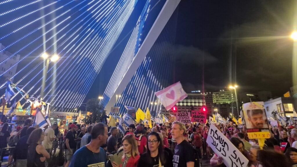 A nighttime photograph capturing a massive public demonstration in Jerusalem beneath the striking blue LED cables of the Chords Bridge, where a dense crowd waves Israeli and yellow hostage flags while carrying signs demanding the release of captives and an end to the Gaza war, visually embodying the societal rupture and civic unrest that characterize Israel's New Exodus, a mass movement exposing deep failures within the ruling coalition.