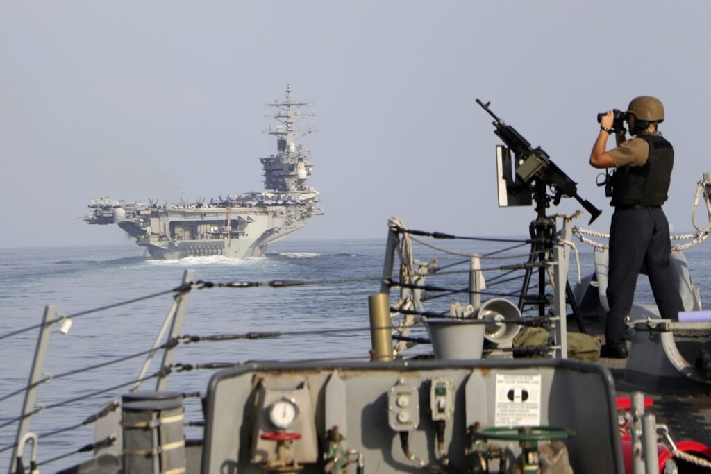 Foreground view from a US Navy warship showing a sailor in tactical gear observing the horizon through binoculars while standing near a mounted machine gun, with a large aircraft carrier navigating the open waters of the Strait of Hormuz in the distance. The scene vividly encapsulates the persistent military vigilance and strategic standoff characterizing the US Naval Blockade of Iran—a high-stakes geopolitical maneuver that has successfully purchased strategic time but ultimately failed to secure lasting peace in the region.