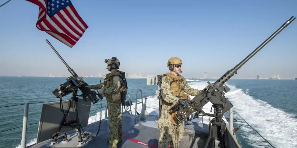 US Pressure on Iran Fails Without Political Deal A high-speed photograph of two US Navy sailors in tactical gear manning heavy deck-mounted machine guns on a fast patrol boat, with the American flag waving against a distant Middle Eastern skyline, visually representing the military component of US Pressure on Iran that ultimately proves ineffective without a complementary political deal.