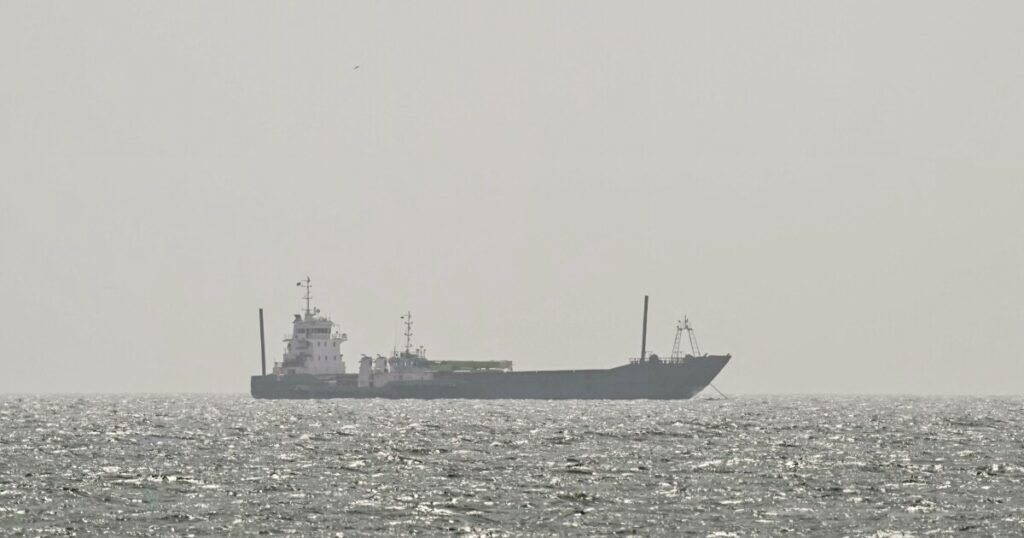 Iran Sanctions Evasion Cripples UAE Trade Ban A distant, hazy photograph of a dark-hulled cargo vessel transiting choppy grey waters, serving as a stark visual metaphor for the shadow fleet operations that underpin Iran Sanctions Evasion and effectively cripple the enforcement of the UAE trade ban.