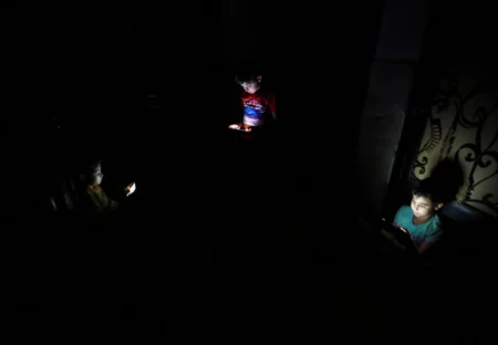 A stark, high-contrast photograph showing three children sitting in pitch darkness, their faces illuminated only by the cold, artificial glow of the smartphones they are holding, vividly capturing the devastating electricity blackouts and daily hardships caused by the Egypt energy crisis that is forcing a desperate green transition amid the ongoing regional war.