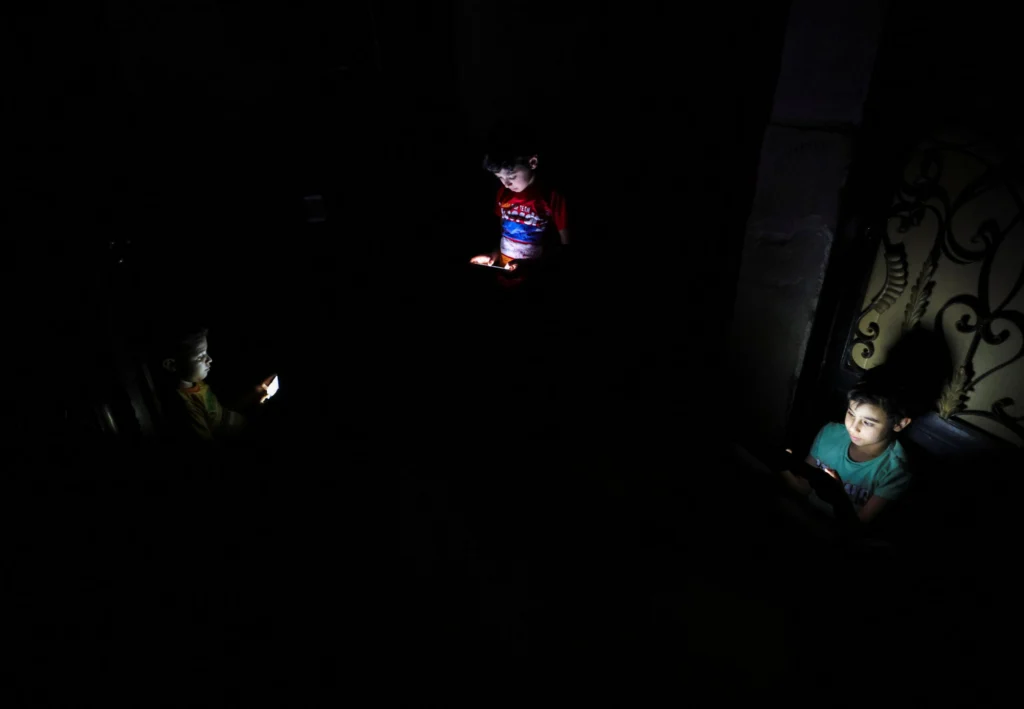 أزمة الطاقة في مصر تجبر على التحول الأخضر وسط الحرب A stark, high-contrast photograph showing three children sitting in pitch darkness, their faces illuminated only by the cold, artificial glow of the smartphones they are holding, vividly capturing the devastating electricity blackouts and daily hardships caused by the Egypt energy crisis that is forcing a desperate green transition amid the ongoing regional war.