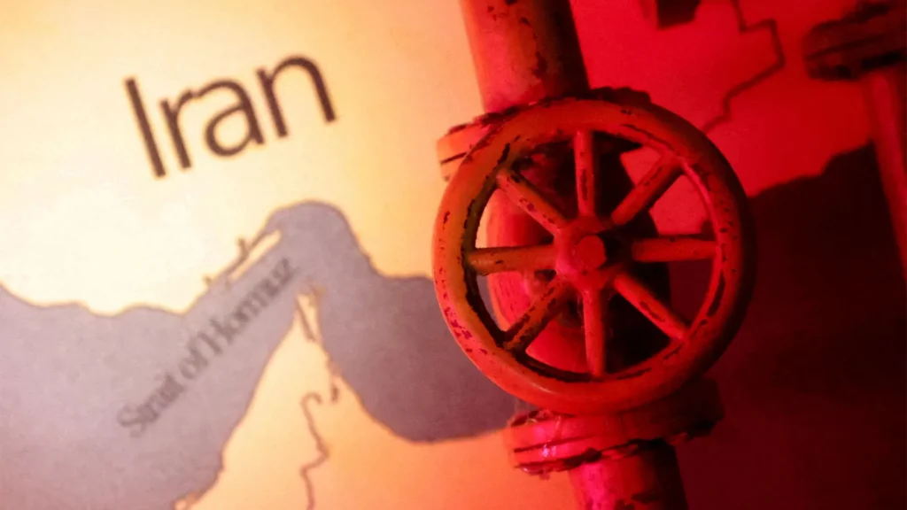 صدمة الطاقة الإيرانية تكشف عن تآكل الاحتياطيات العالمية Close-up of an industrial pipeline valve wheel against a map featuring Iran and the Strait of Hormuz under red lighting, illustrating vulnerabilities driving an Iran Energy Shock.