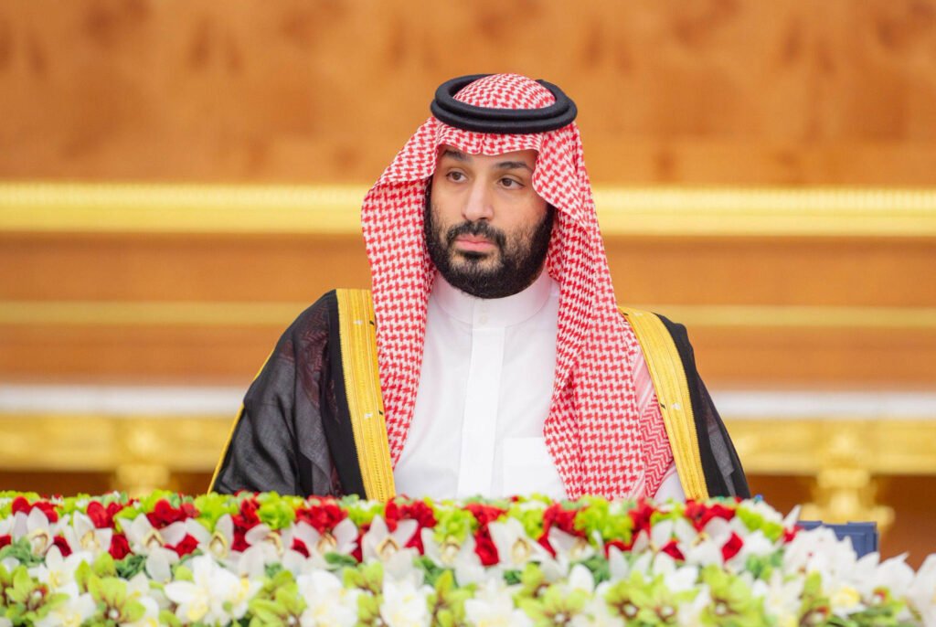 Saudi Assertiveness Counters Iran’s Proxies, Risks War Saudi Crown Prince Mohammed bin Salman sits at a formal podium adorned with flowers, reflecting the leadership posture central to growing Saudi assertiveness.