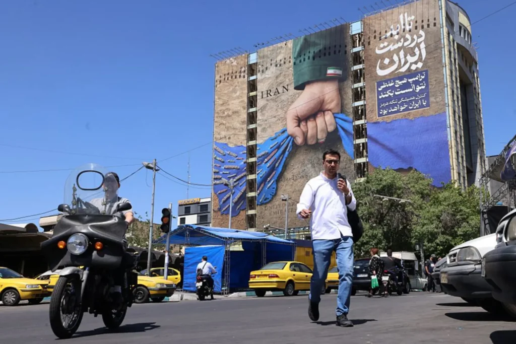 تجنب صدمة النفط العالمية حتى الآن في حرب إيران A street-level photograph of a man crossing Vanak Square in Tehran, with a motorcyclist and yellow taxis passing by, set against a massive billboard on a building depicting a fist holding the blue map of the Strait of Hormuz with the word "IRAN", symbolizing Tehran's strategic maritime leverage that has so far prevented a major Global Oil Shock during the Iran war.