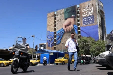 A street-level photograph of a man crossing Vanak Square in Tehran, with a motorcyclist and yellow taxis passing by, set against a massive billboard on a building depicting a fist holding the blue map of the Strait of Hormuz with the word "IRAN", symbolizing Tehran's strategic maritime leverage that has so far prevented a major Global Oil Shock during the Iran war.