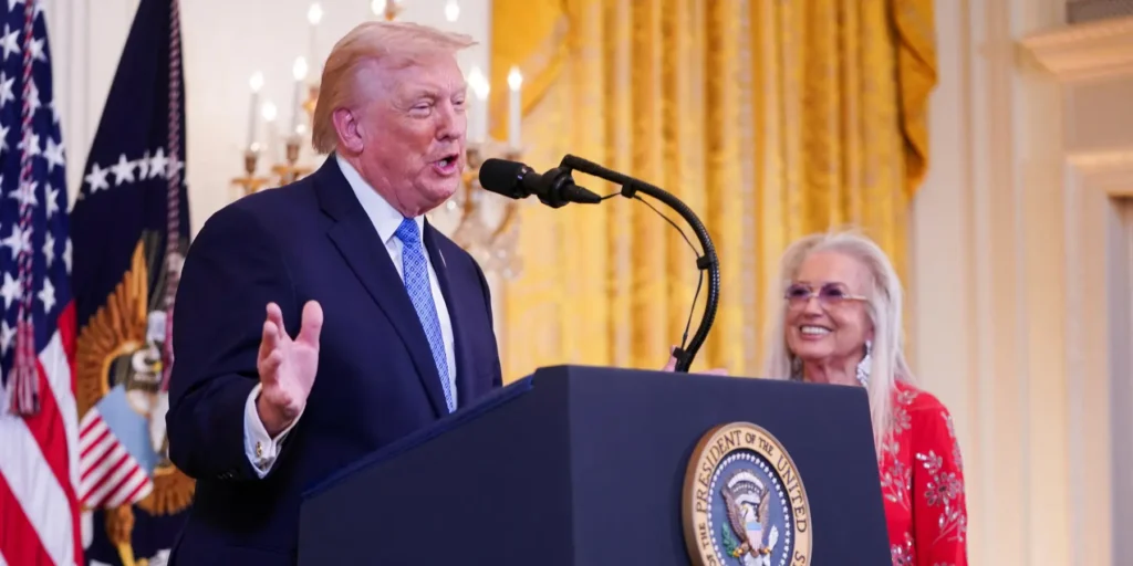 Israel Lobby Influence Distorts American Elections A photograph of President Donald Trump speaking at the White House podium, with prominent pro-Israel donor Miriam Adelson smiling behind him in a red dress, visually illustrating the deep political entanglement and the powerful Israel Lobby Influence that critics argue distorts American elections.