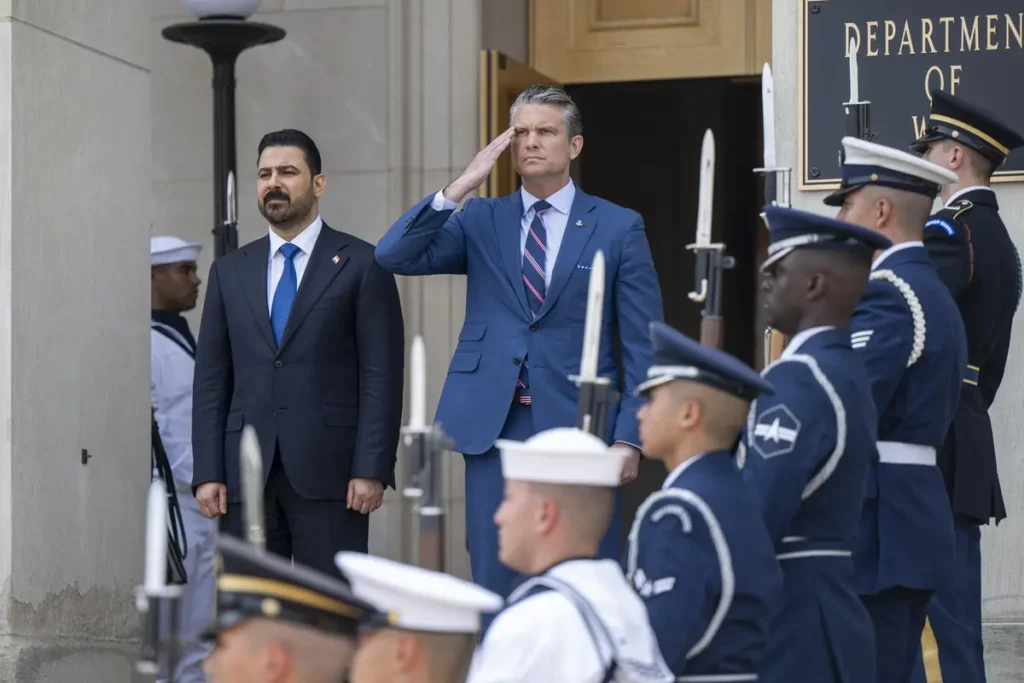 تعارض خلع سلاح شبهنظامیان عراقی با فشارهای ایرانی A formal diplomatic photograph showing US Secretary of Defense Pete Hegseth saluting alongside Iraqi Defense Minister Thabet al-Abassi outside the Department of Defense entrance, flanked by a ceremonial military honor guard with bayonets, illustrating the high-stakes bilateral negotiations where Iraqi Militia Disarmament collides with severe Iranian political and military pressure.