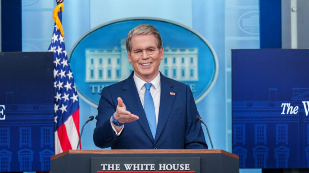 A bright photograph of Treasury Secretary Scott Bessent in a navy blue suit smiling and gesturing from the White House press briefing podium, flanked by US flags and the official seal, visually representing the administration's public response to the escalating Treasury Bond Blues as investor alienation and desperate buyback measures signal a collapsing market.