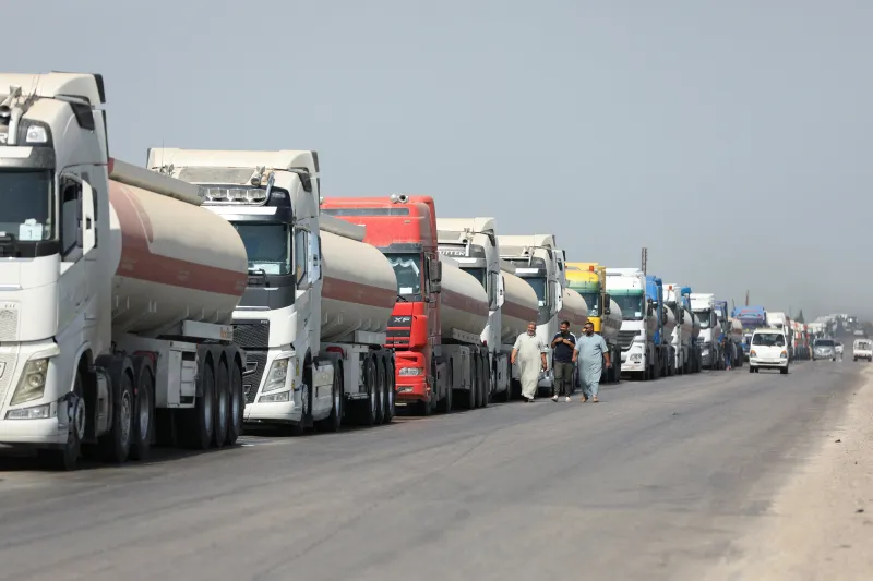 ممرات التجارة في سوريا تدفع نحو انتعاش محفوف بالمخاطر بعد الأسد A long line of heavy fuel tanker trucks carrying oil along a dusty highway, with men in traditional clothing walking alongside, symbolizing the cross-border convoy from Iraq to the Baniyas terminal that sustains the Syria Trade Corridors, driving the fragile and risky post-Assad recovery in the region.