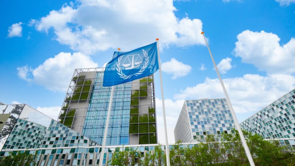 A low-angle photograph showing the bright blue flag of the International Criminal Court (ICC) waving against a cloudy blue sky, set against modern glass buildings with vertical green garden walls in The Hague, symbolizing the international legal institution that remains standing amid escalating political attacks, illustrating how the War on ICC backfires on the United States as it dismantles its own accountability structures.