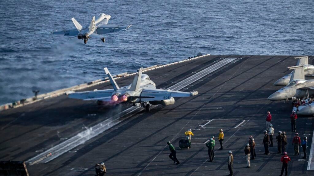 تآكل التفوق العسكري الأمريكي مع استنزاف الحرب مع إيران للاحتياطيات A dynamic photograph of US Navy F/A-18 fighter jets conducting high-tempo launch operations from an aircraft carrier deck with glowing afterburners, surrounded by deck crew in colored vests over the open ocean, illustrating the intense operational strain that threatens to erode the US Military Edge as the prolonged Iran war depletes critical reserves.