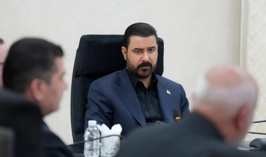 Iran-backed Iraqi militias face pressure after 966 attacks Iraqi Prime Minister Ali Al Zaidi seated at a formal conference table, wearing a navy suit and black shirt with a serious expression, reflecting the high-level political scrutiny and growing government pressure currently facing Iran-backed Iraqi militias following the 966 attacks.