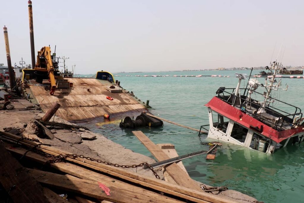 Yemen’s War Reignites as Houthi Attacks Spread A scene at the Port of Mokha showing the sunken red-and-white navigation bridge of a vessel partially protruding above the teal water, surrounded by broken wooden planks, chains, and a floating barge with a yellow excavator, illustrating the maritime devastation inflicted by Houthi strikes that are actively spreading hostilities and reigniting Yemen's War along the Red Sea coastline.