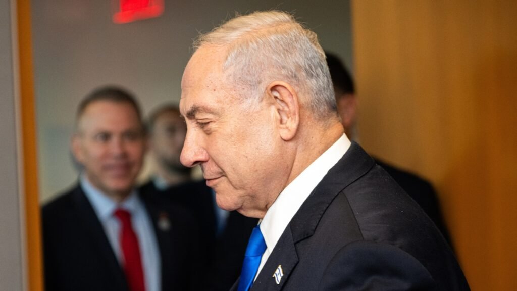 A close-up profile photograph of Israeli Prime Minister Benjamin Netanyahu wearing a dark suit and bright blue tie, captured in a candid moment with a serious expression as he walks past blurred officials under a red exit sign, symbolizing the politically charged decisions surrounding the Israeli Budget that funds West Bank settlements and enforces Haredi draft exemptions.