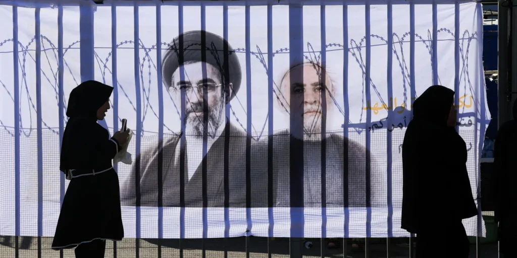 Two silhouetted women walk past a banner depicting Resistance Axis leadership behind vertical bars, illustrating the political skepticism in Beirut surrounding prospective Trump-Hezbollah Talks.