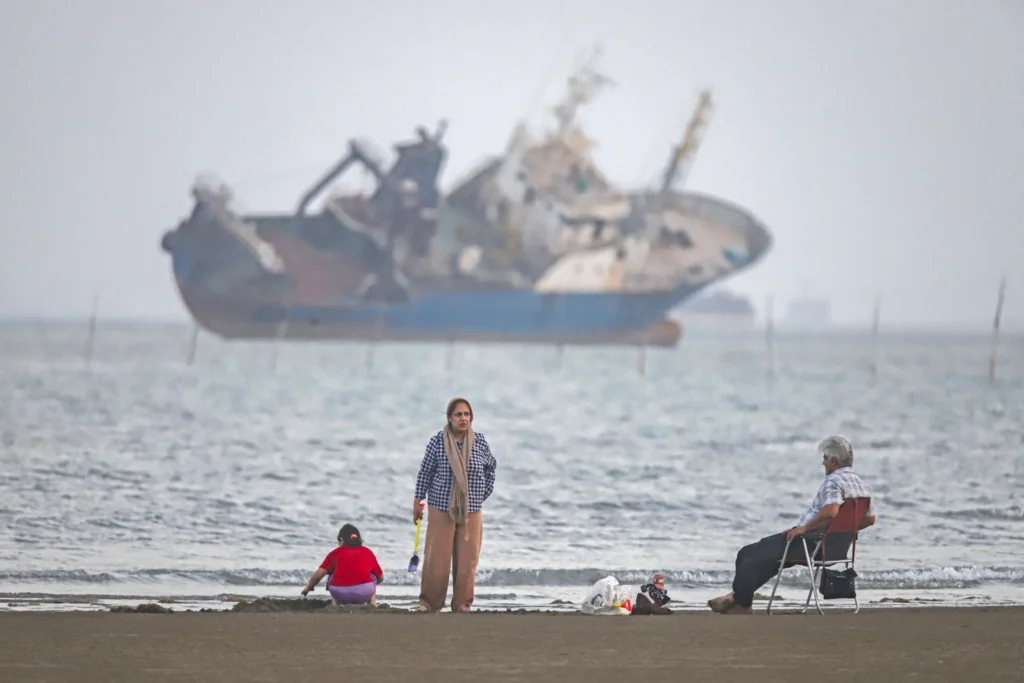 Iran Economic Options: Resolving the Hormuz Dilemma "A hazy coastal scene showing civilians relaxing on a beach—a standing woman, a child playing in the sand, and an elderly man seated in a folding chair—contrasted sharply with a massive, rusted, and partially sunken shipwreck looming in the foggy offshore waters, serving as a stark visual metaphor for the maritime insecurity in the Strait of Hormuz that directly restricts Iran Economic Options and underscores the urgent need for a diplomatic resolution to restore regional trade stability."
