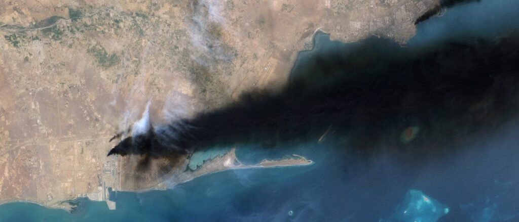 A stark satellite image showing a massive, dark plume of smoke rising from a coastal infrastructure site along an arid shoreline, with the thick black cloud sweeping forcefully over the blue Persian Gulf waters, symbolizing the destructive physical footprint of the Houthi Escalation that is widening the regional conflict and placing Saudi Arabian energy and security infrastructure directly in the crosshairs.