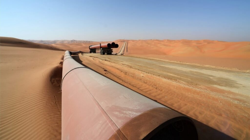 Regional Energy Routes Strengthened by Mecca Pact A wide-angle desert photograph showing a large, reddish-brown pipeline stretching across rippled orange sand dunes, while a flatbed truck transports another section of the pipe along a dirt road, symbolizing the physical infrastructure and development driving the strengthened Regional Energy Routes established by the Mecca Pact.