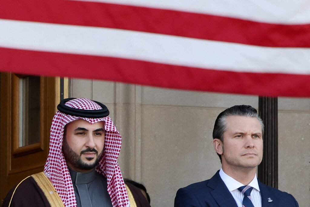 إعادة التفكير الاستراتيجي في القواعد الأمريكية بالخليج بعد الحرب الإيرانية "U.S. Defense Secretary Pete Hegseth stands alongside Saudi Defense Minister Prince Khalid bin Salman, who wears traditional red-and-white headwear and a dark bisht with gold trim, underneath the striped American flag at the Pentagon, capturing the high-level diplomatic engagement that will govern the security arrangements and operational status of US Bases in Gulf as regional military strategies shift in the aftermath of the Iran war."