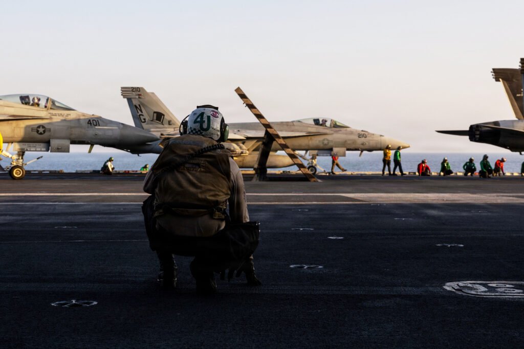 نهاية اللعبة الإيرانية تتطلب فتح مضيق هرمز وإزالة الأسلحة النووية A strategic photograph of a US Navy flight deck crew member in protective gear crouching on the carrier deck, observing F/A-18 fighter jets and other sailors preparing for launch operations against a low-lit ocean horizon, visually representing the naval military readiness driving the Iran Endgame demands for a permanently open Strait of Hormuz and full nuclear removal.