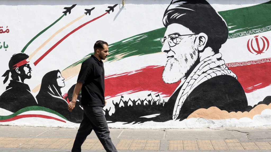 ایران پس از جنگ به دنبال نقشی در نظم جدید خاورمیانه است A street-level photograph showing a man in a black polo shirt walking past a large Tehran mural featuring a portrait of Ayatollah Ali Khamenei, fighter jets, revolutionary silhouettes, and the colors of the Iranian flag, representing the enduring ideological foundation and military symbolism that define Postwar Iran as it maneuvers for a leading strategic role in the new Middle East order.