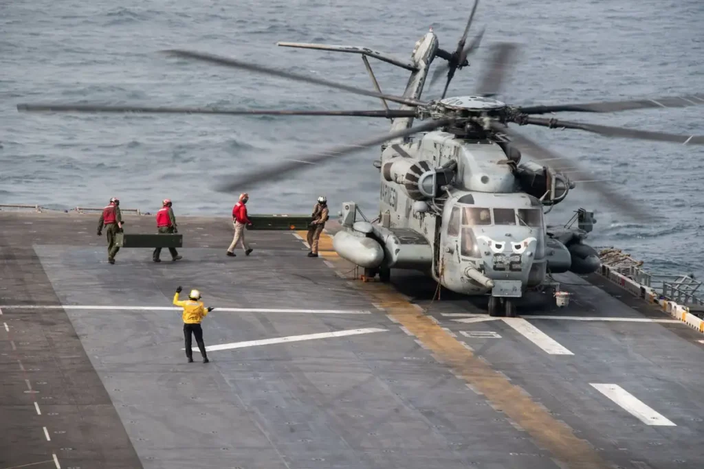 Iran’s Hormuz Leverage Collapses as Blockade Fails A wide shot of a grey military transport helicopter with blurred spinning rotors resting on the flight deck of a naval warship on the open ocean, while deck crew members in high-visibility red and yellow vests conduct flight line operations, symbolizing the unyielding international naval presence that has successfully neutralized Tehran's maritime threats, causing Hormuz Leverage to collapse as the long-projected blockade fails to interdict global shipping lanes.