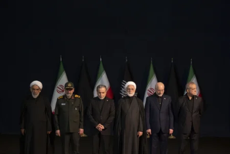 A solemn group photograph of Iran's top civilian and military leadership standing in a unified line against a dark backdrop of Iranian and black mourning flags, attending a high-level state funeral, visually portraying the enduring and unified Iranian Regime Resilience that continues to defy external maximum pressure campaigns.