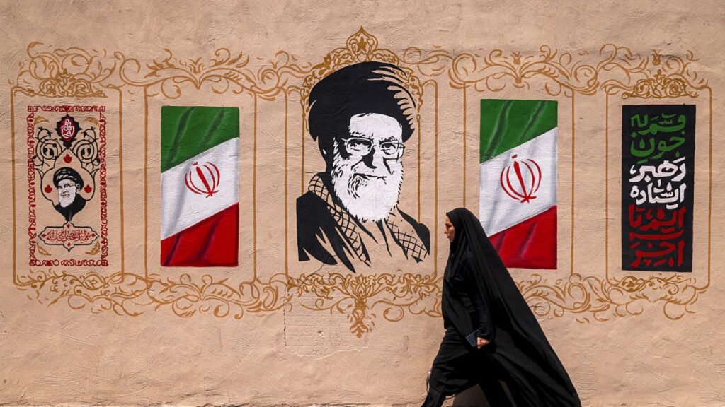 A woman in a chador walks past a street mural featuring Iranian Supreme Leader Ali Khamenei and state symbols, illustrating the ideological backdrop driving Iran factional politics.