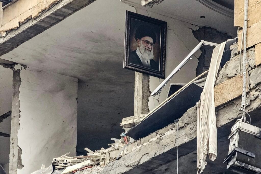 Iran War Fallout Forces US Middle East Reckoning A photograph of a framed portrait of Iran's late Supreme Leader Ayatollah Ali Khamenei hanging precariously on the exposed, shattered concrete wall of a destroyed building in Haret Hreik, Lebanon, surrounded by tangled debris and broken window frames, symbolizing the widespread devastation and regional instability that constitutes the Iran War fallout, which now forces a critical US Middle East reckoning.