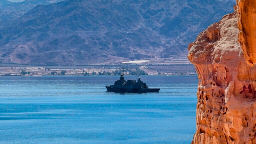 A grey naval warship navigating through deep blue waters, framed by a rugged, orange rock cliff and barren desert mountains in the background, illustrating the operational presence of the Saudi Maritime Coalition, which analysts highlight as a clear signal of the declining US security umbrella in the region.
