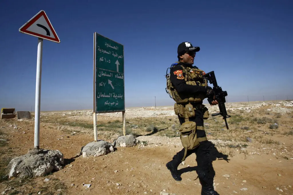 Saudi Doctrine on Iraq: Can Bombs Back the State? An armed Iraqi security officer in tactical gear patrols a desert road beside a green sign marking the Iraqi-Saudi border 105 km away, illustrating the Saudi Doctrine on Iraq and whether military force can back the state.