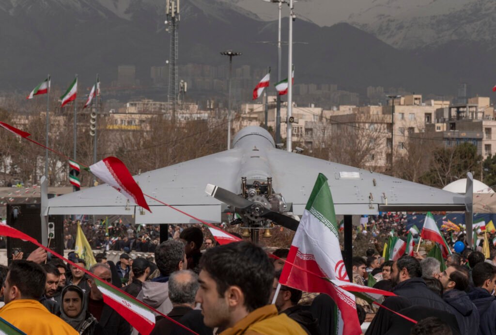 A grey Shahed drone with its propeller visible sits on a display platform above a crowd waving Iranian flags, with snow-capped mountains behind Tehran's skyline — a public display embodying the Iran Deterrence doctrine as it shifts from patience to counter-escalation.