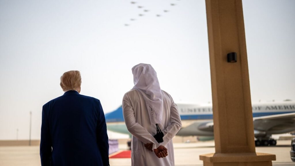 چرا شراکت خلیج-آمریکا در جنگ ایران ادامه خواهد یافت A photograph showing the backs of a US President in a dark suit and a Gulf Arab leader in traditional white attire standing side-by-side on an airfield tarmac, looking up at a formation of military jets flying overhead with Air Force One in the hazy background, symbolizing the deep strategic and defense coordination that ensures the Gulf-US Partnership will endure beyond the Iran war.