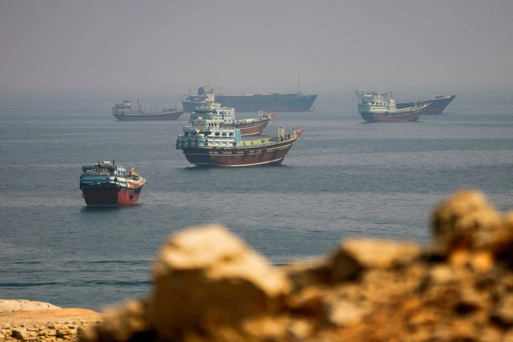 Gulf Maritime Mechanism: A Face-Saving Exit for Iran Wooden cargo dhows anchor in a hazy Gulf seascape viewed through blurred rocks, illustrating the Gulf Maritime Mechanism that offers Iran a face-saving exit.
