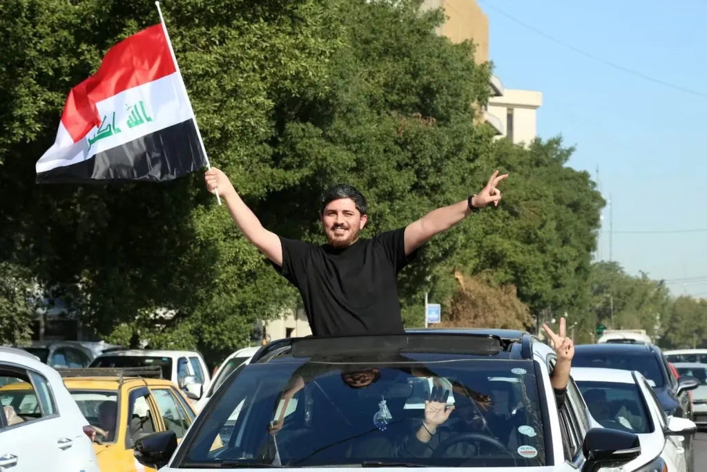 آزادی مطبوعات در عراق، انحصار دولتی را به چندین ارباب رقابتی واگذار کرد A smiling man stands through the sunroof of his car, waving the Iraqi national flag and flashing a peace sign above a crowded Baghdad street, a scene of public expression that sits at the heart of the debate over Iraq Press Freedom.