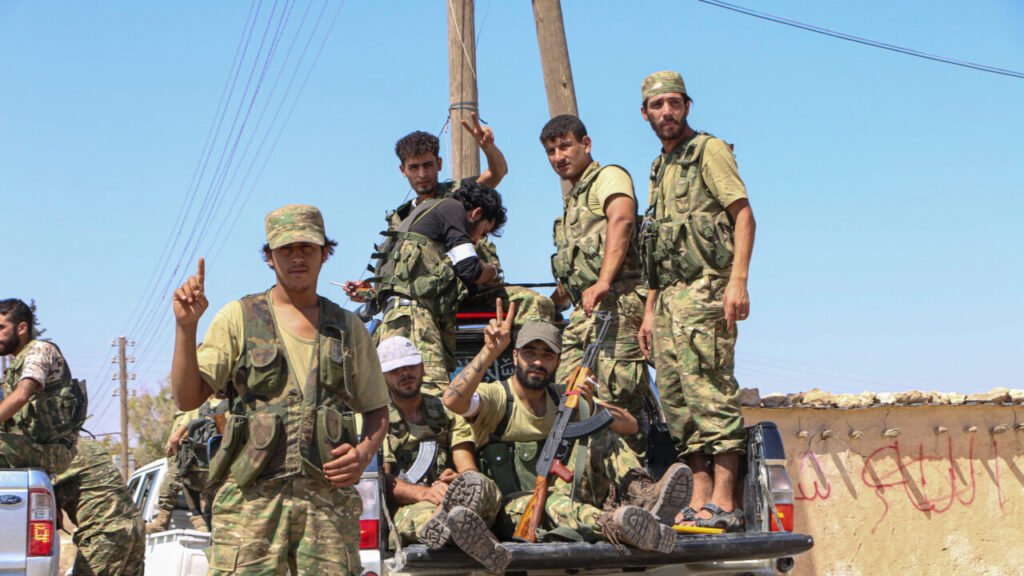 A photograph of soldiers in camouflage fatigues celebrating on the back of a pickup truck in Aleppo, making V-signs and holding rifles after defeating ISIS in 2017, capturing the historical zenith of the Syrian Democratic Forces before their recent integration into the Damascus regime, a transition that now offers enduring strategic lessons for the United States.