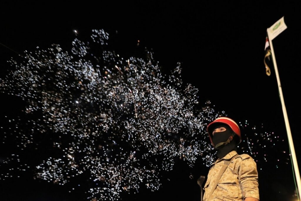 A Houthi security guard in desert camouflage and a black face covering stands beneath a night sky bursting with fireworks during a ceremony in Sanaa marking the 12th anniversary of the capital's takeover, a moment of defiant display as US-Houthi Talks test whether Iran wears Washington down.