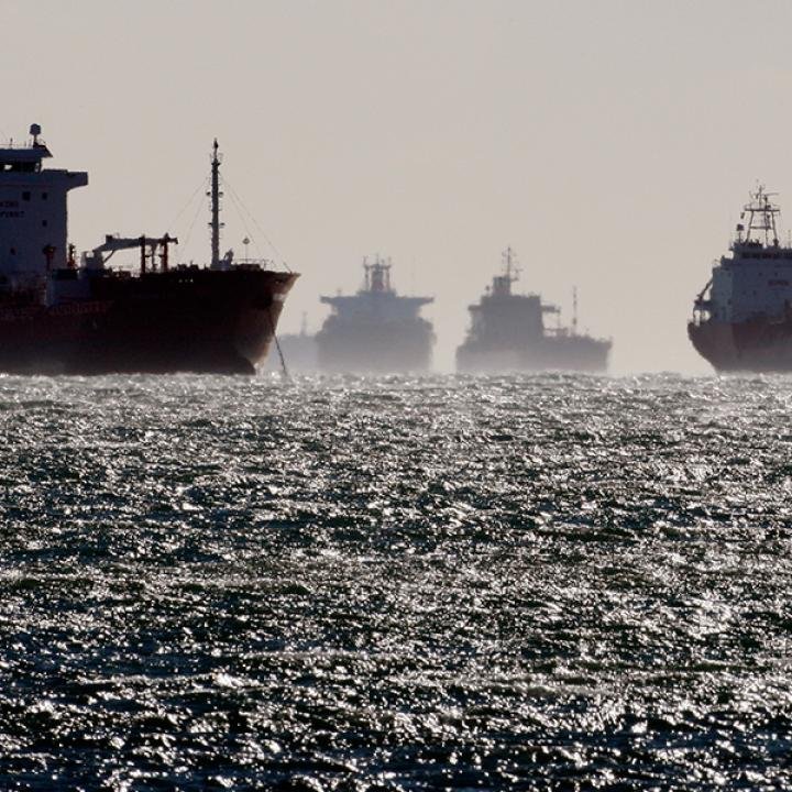 Hormuz Shuttle Tankers Sail On After Deadly Iranian Hit Several large tankers sit silhouetted in hazy, sunlit waters, their dark hulls receding into the distance as light scatters across the choppy sea — the scene of Hormuz Shuttle Tankers still sailing on after a deadly Iranian hit.